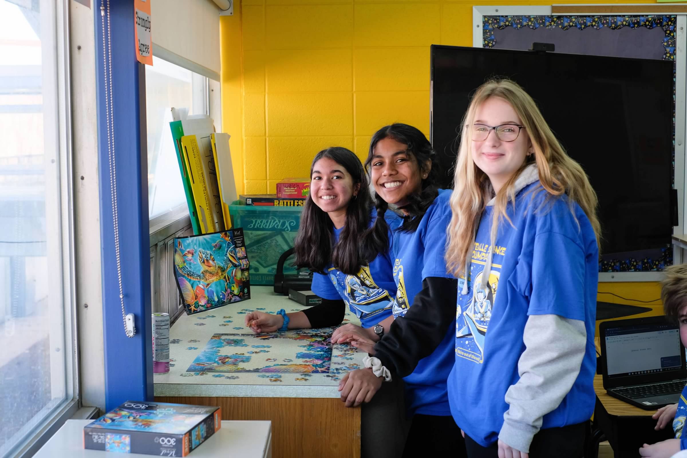 3 students smiling while standing over unfinished puzzle on table